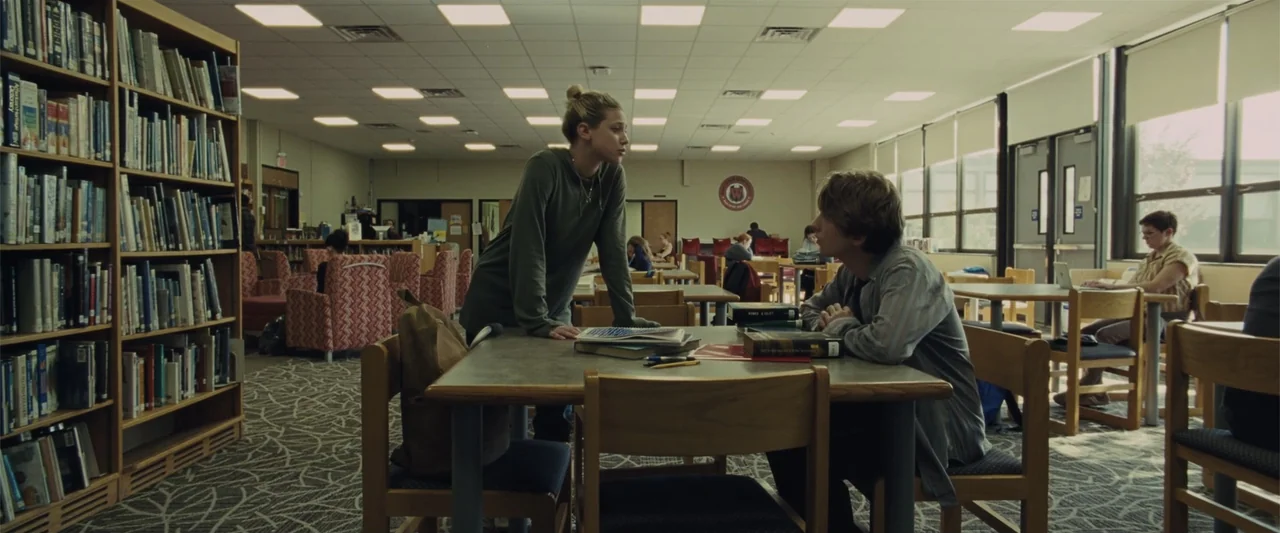 Chemical Hearts (2020) movie still: medium — A quiet school library with rows of bookshelves and rows of tables with chairs. A female student stands…