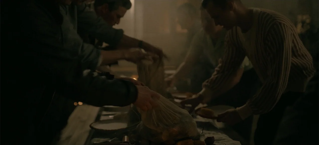 Devotion (2022) movie still: medium — A dimly lit interior room contains a long wooden table with plates and food. Several men stand around the…