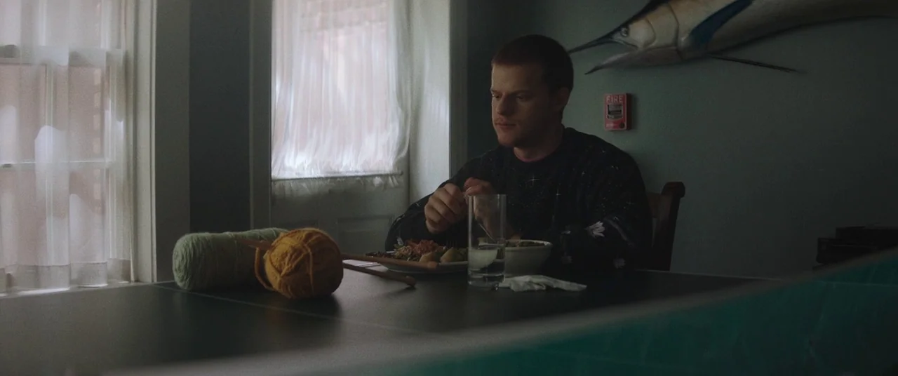 Honey Boy (2019) movie still: medium — A man sits at a dining table beside a large wall-mounted decorative marlin trophy. He eats his meal while a…