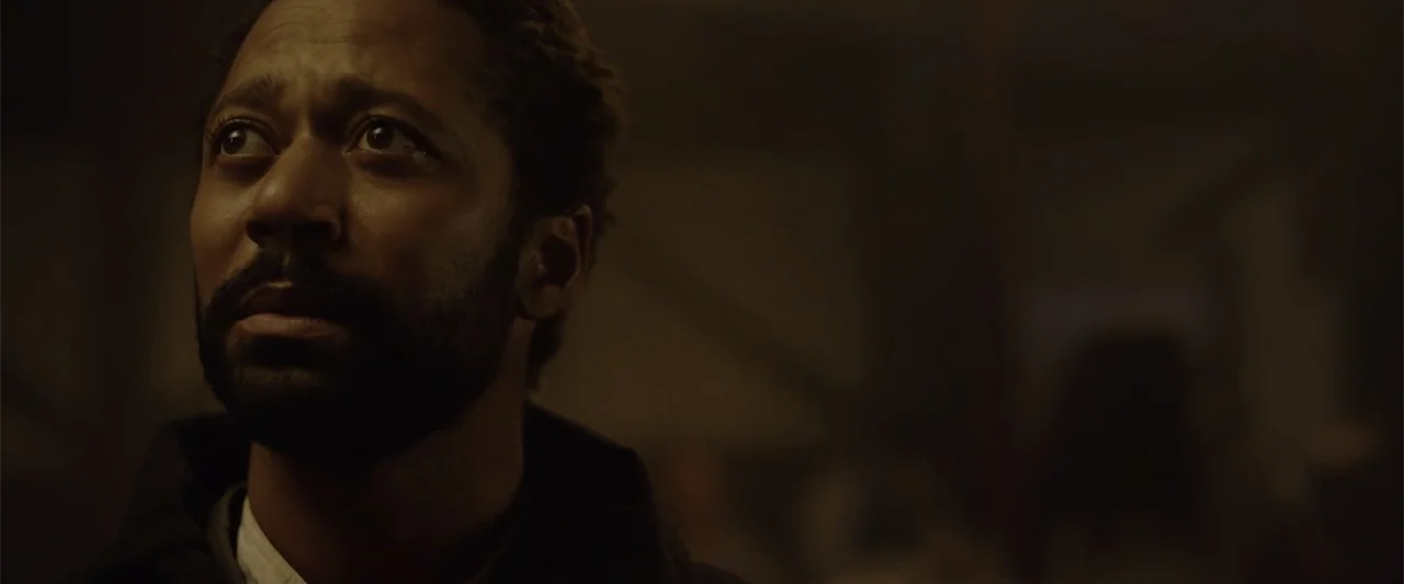 Ghosts of the Ozarks (2022) movie still: closeup — A dimly lit, shadowed industrial space with indistinct structures in the background. A man with a…