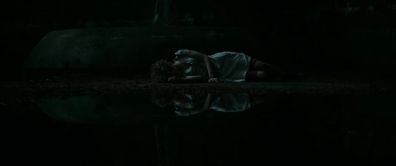 It (2017) movie still: wide — A dark outdoor area features a large, still puddle reflecting a reclining figure. The woman rests on the damp ground…