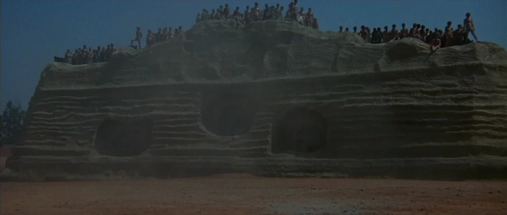 Fellini – Satyricon (1969) movie still: wide — A massive, sculpted rock formation sits on a dusty, flat, reddish-brown earthen expanse. A large…