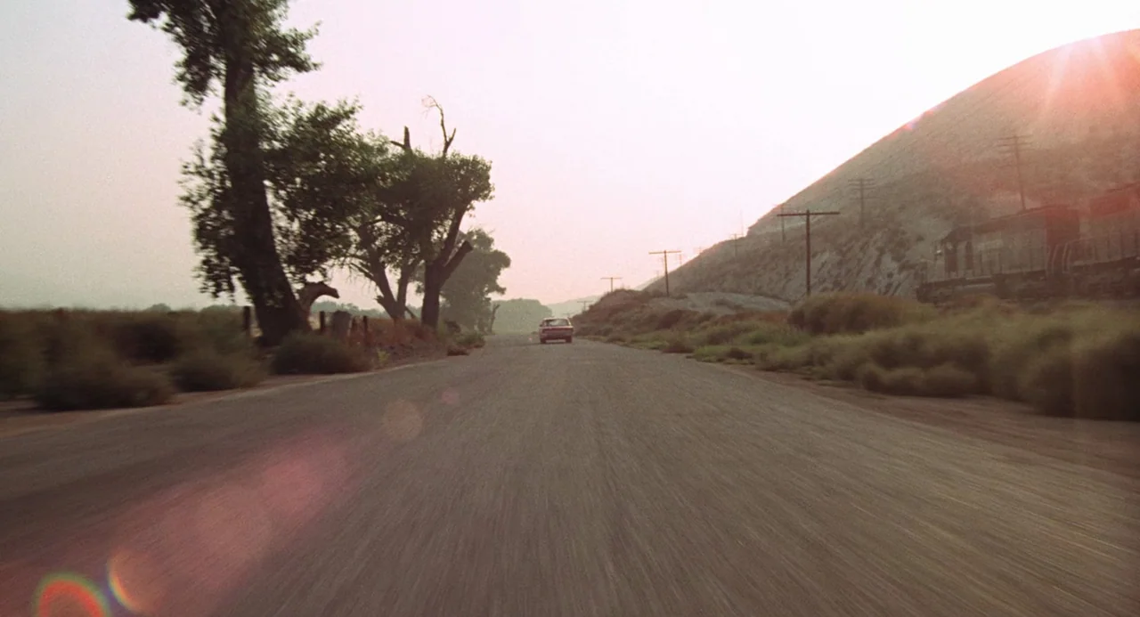 Duel (1971) movie still: wide — A rural paved road stretches toward a distant horizon flanked by trees and hills. A single vintage sedan drives away…