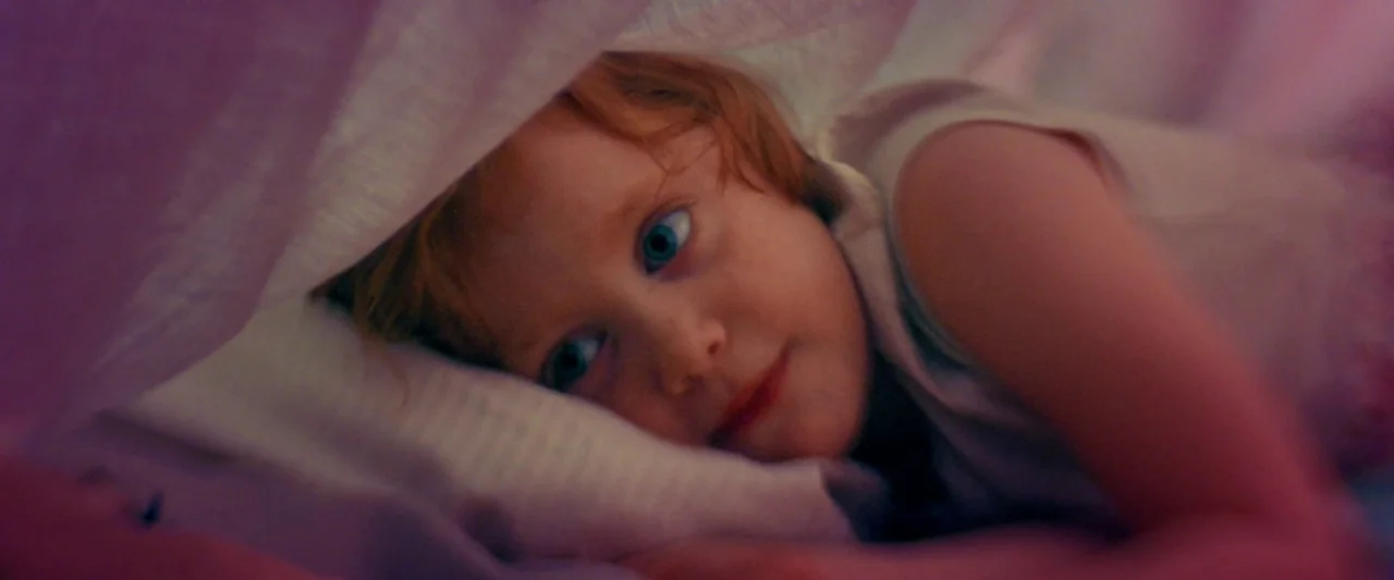 Moon Garden (2022) movie still: closeup — A child rests beneath a soft pink fabric layer in a dark environment. The child gazes outward from their…