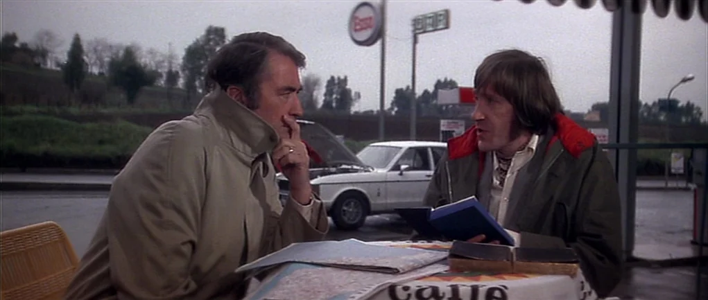 The Omen (1976) movie still: medium — An outdoor gas station forecourt during an overcast day with a car in background. Two men sit at a round table…