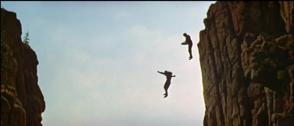 A deep gorge separates two towering rock walls under a bright daylight sky. Two men are captured in mid-air as they fall between the cliffs.