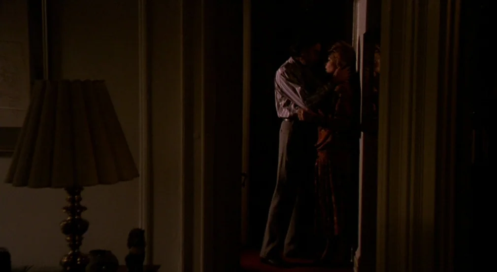 Crimes and Misdemeanors (1989) movie still: medium — A dimly lit doorway connects two dark rooms in a house interior. A man and a woman stand within…