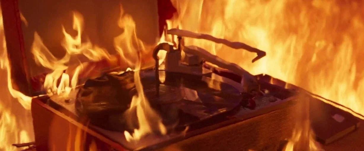 Last Night in Soho (2021) movie still: closeup — A wooden record player is surrounded by high, licking flames in a dark space. The vinyl disc sits…