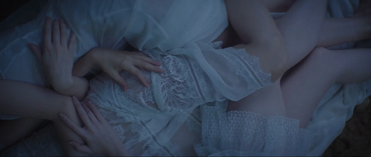 Braid (2018) movie still: closeup — Two individuals dressed in white lace clothing lie together on a white sheet. They are wrapped around each other…