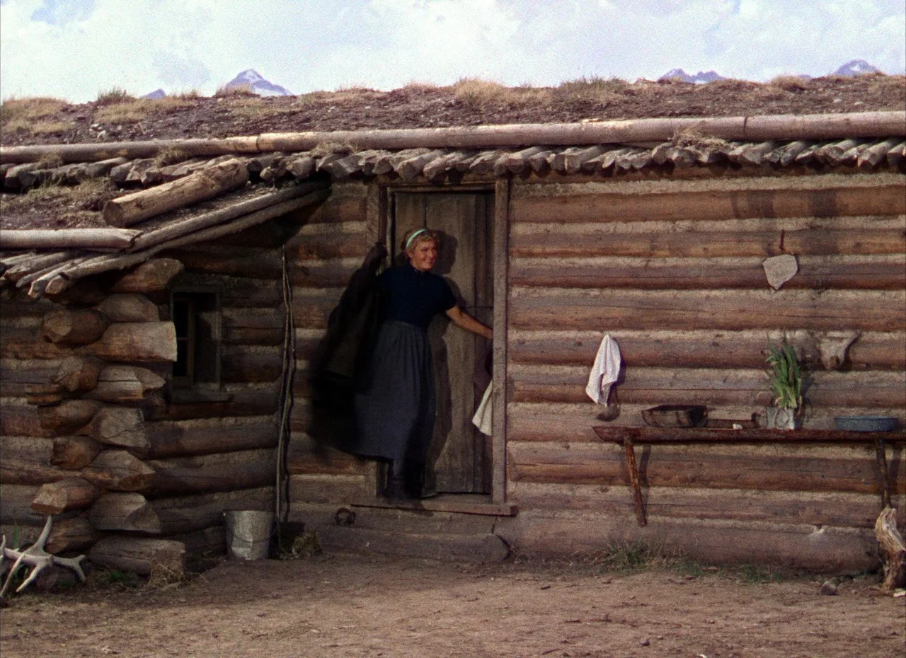 Shane (1953) movie still: fullbody — A rustic log cabin with a sod roof stands against a mountain backdrop. A woman exits the cabin door while…