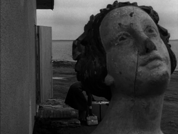 Persona (1966) movie still: closeup — A stone sculpture head sits in the foreground beside a coastal building. A person is visible in the background…