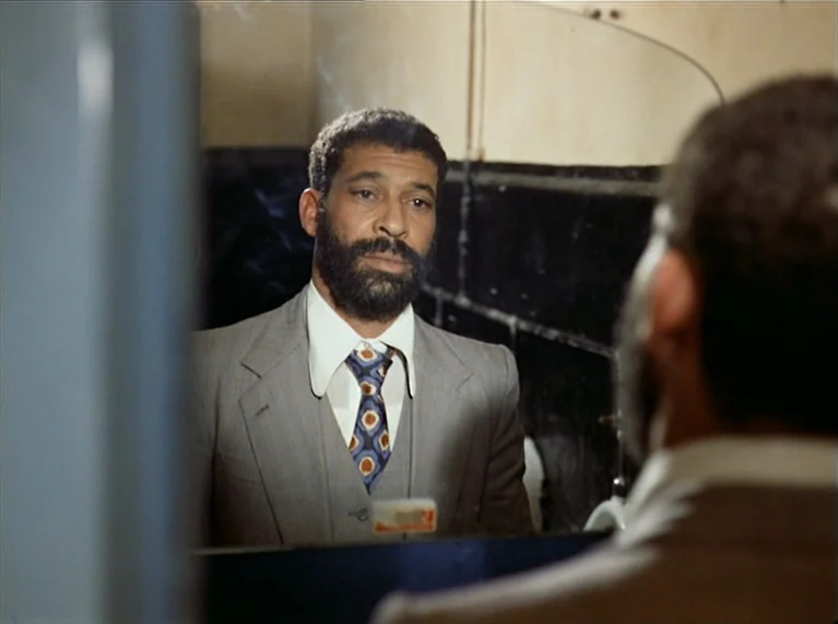 Ali: Fear Eats The Soul (1974) movie still: medium — A public restroom interior features black wall tiling and a rectangular wall mirror. A man…