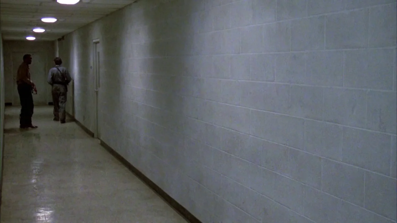 Day of the Dead (1985) movie still: wide — A long, narrow hospital corridor features pale white block walls and tiled flooring. Two men stand…
