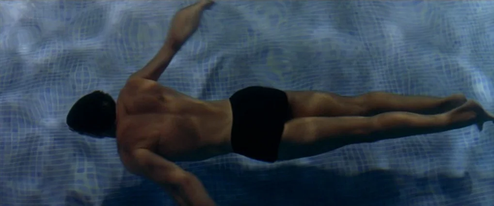 Talk To Her (2002) movie still: fullbody — A man swims underwater in a pool with a tiled blue grid floor. The sun illuminates the water creating…