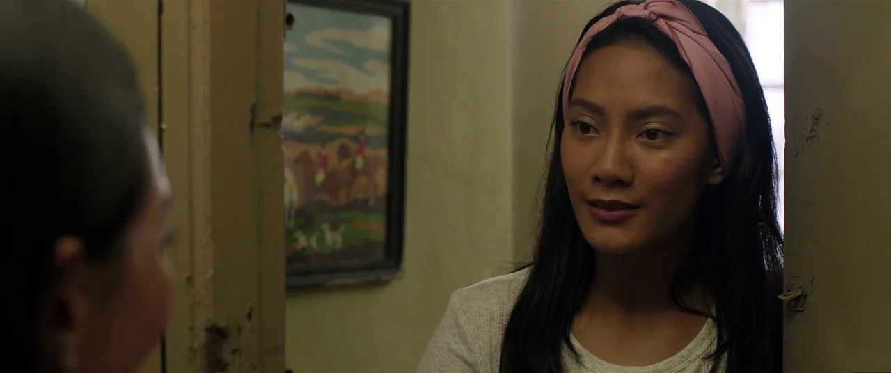 Satan’s Slaves (2017) movie still: medium — A dimly lit doorway separates two areas in a home with neutral-colored walls. A young woman with a pink…