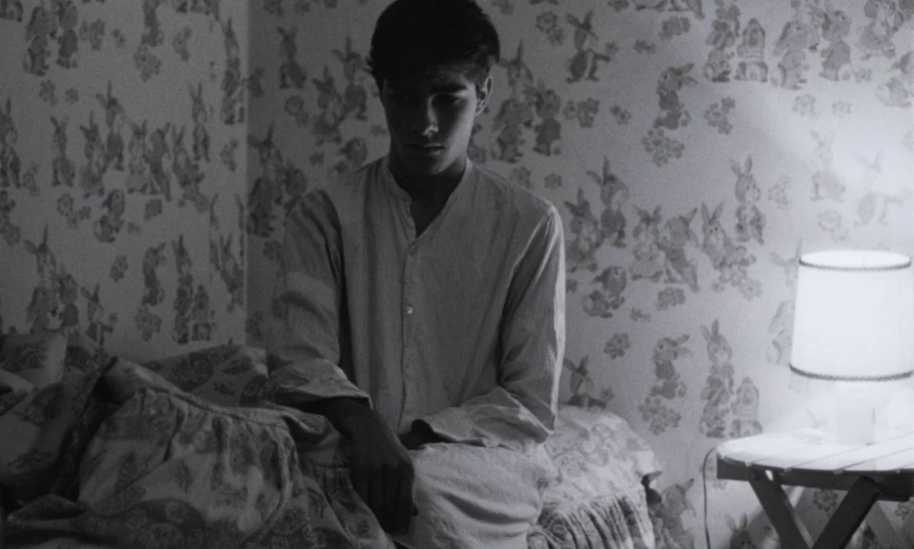 The Secret Son (1979) movie still: medium — A bedroom wall features wallpaper with a repeating rabbit pattern. A young male sits on the edge of the…