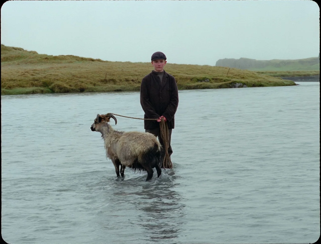 Godland (2022) movie still: fullbody — A wide, shallow river flows through a grassy, undulating landscape under an overcast sky. A young boy in…