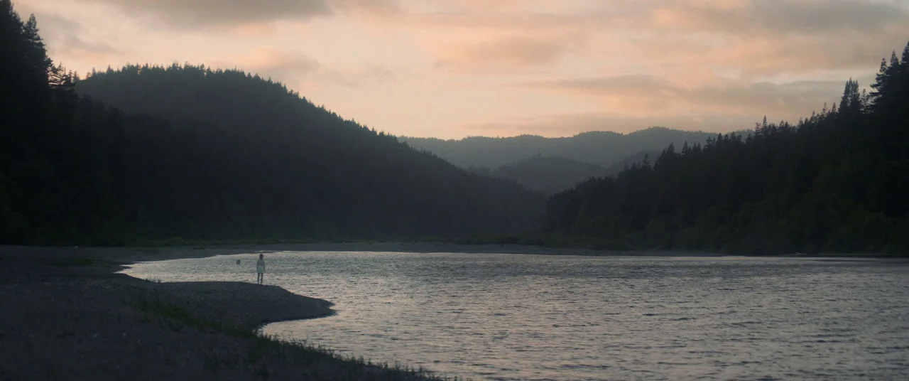 Woodshock (2017) movie still: wide — A wide river winds between dense coniferous forests and dark mountain slopes at sunset. A lone figure in a…