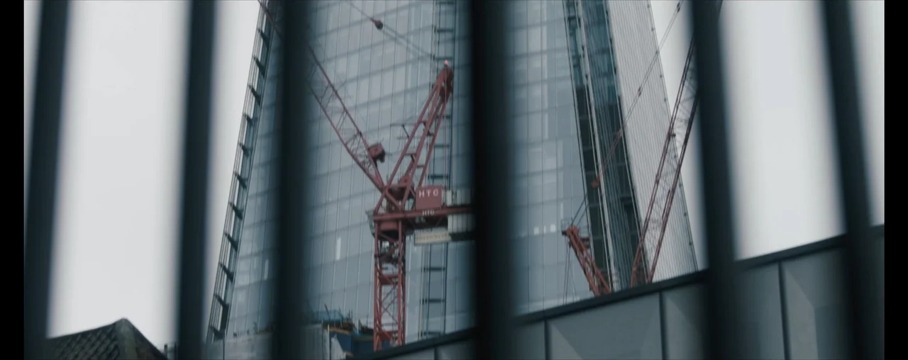 The Wrong Tenant (2022) movie still: wide — A glass skyscraper under construction is viewed through a foreground of vertical steel bars. A large red…