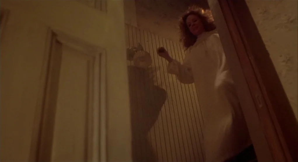 Carrie (1976) movie still: fullbody — An interior wooden staircase features pale vertical-patterned wallpaper and a dimly lit fixture. A woman in a…