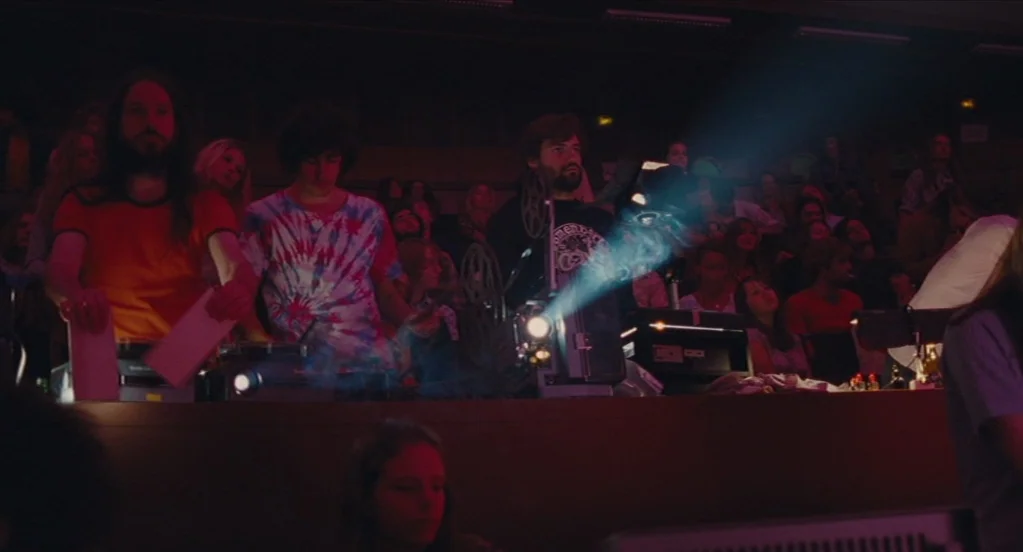 Something in the Air (2012) movie still: medium — A dark, crowded projection booth with three men operating 16mm film projectors. They watch as the…