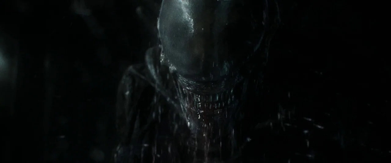 Alien: Covenant (2017) movie still: closeup — A dark, textured alien creature with a smooth, elongated cranium dominates the dark frame. Viscous…