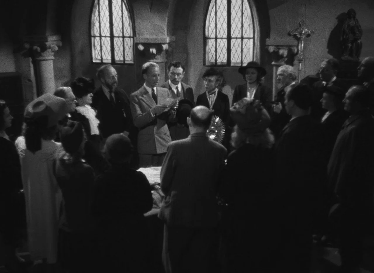 Le Corbeau (1943) movie still: wide — A stone interior with arched, leaded-glass windows and a religious crucifix on the wall. A group of men and…