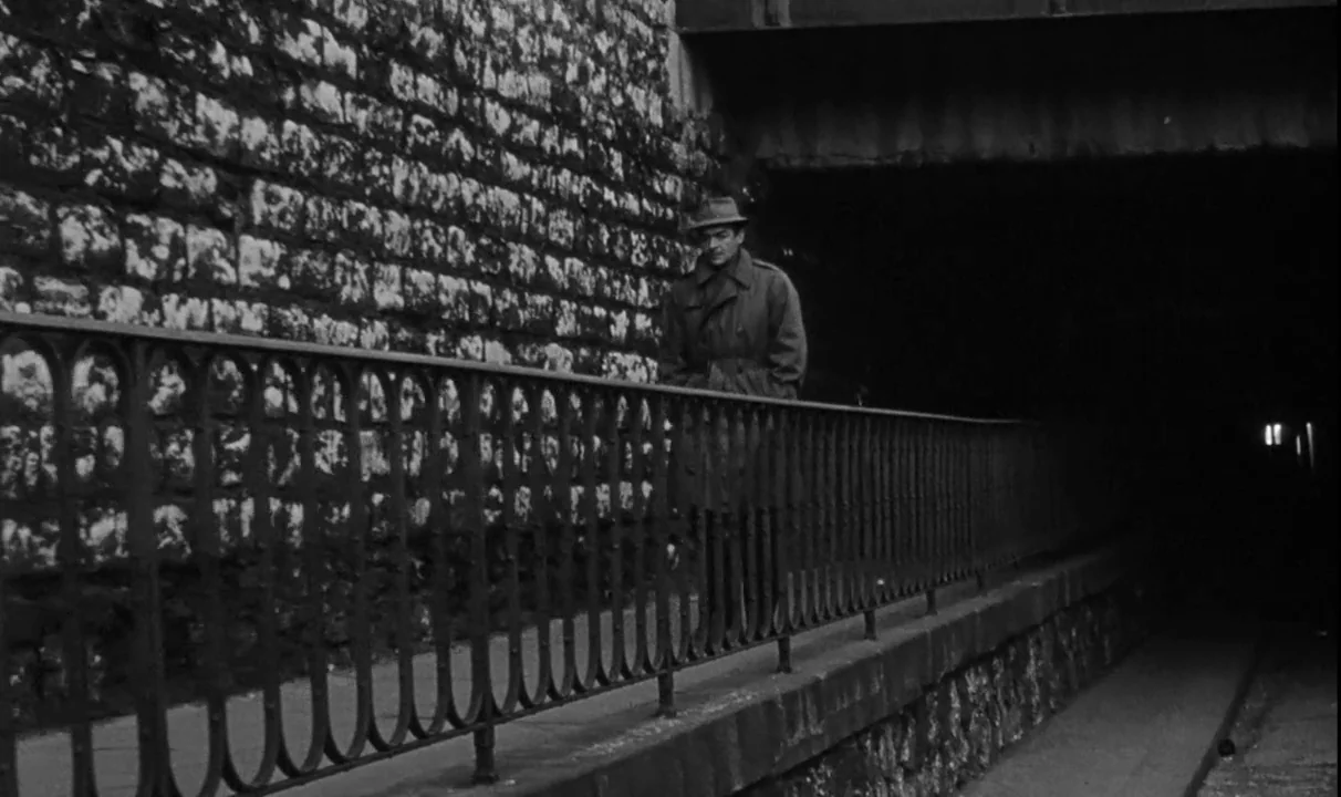 Le Doulos (1962) movie still: fullbody — A stone wall runs parallel to an elevated metal railing by a dark tunnel. A man in a trench coat and fedora…
