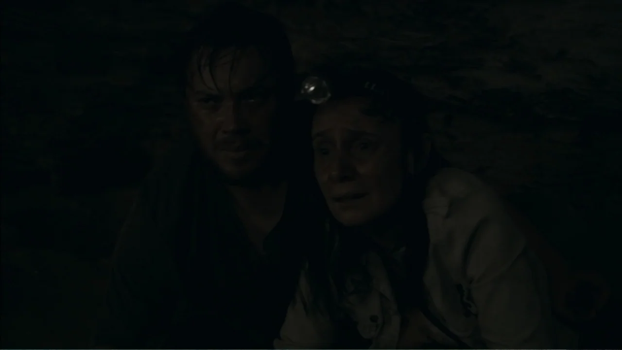 The Grotto (2022) movie still: medium — A dark, cramped stone environment with jagged rock textures visible in the background shadows. Two people…