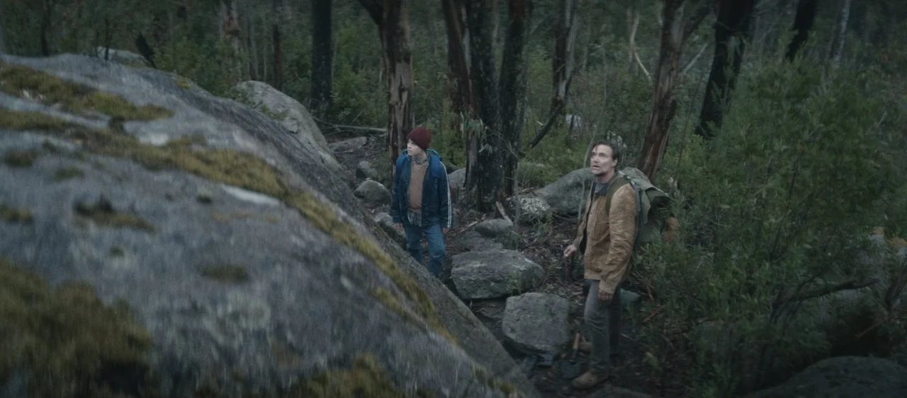 Force of Nature: The Dry 2 (2024) movie still: fullbody — A dense forest path with large granite boulders and tall, sparse trees. Two people with…