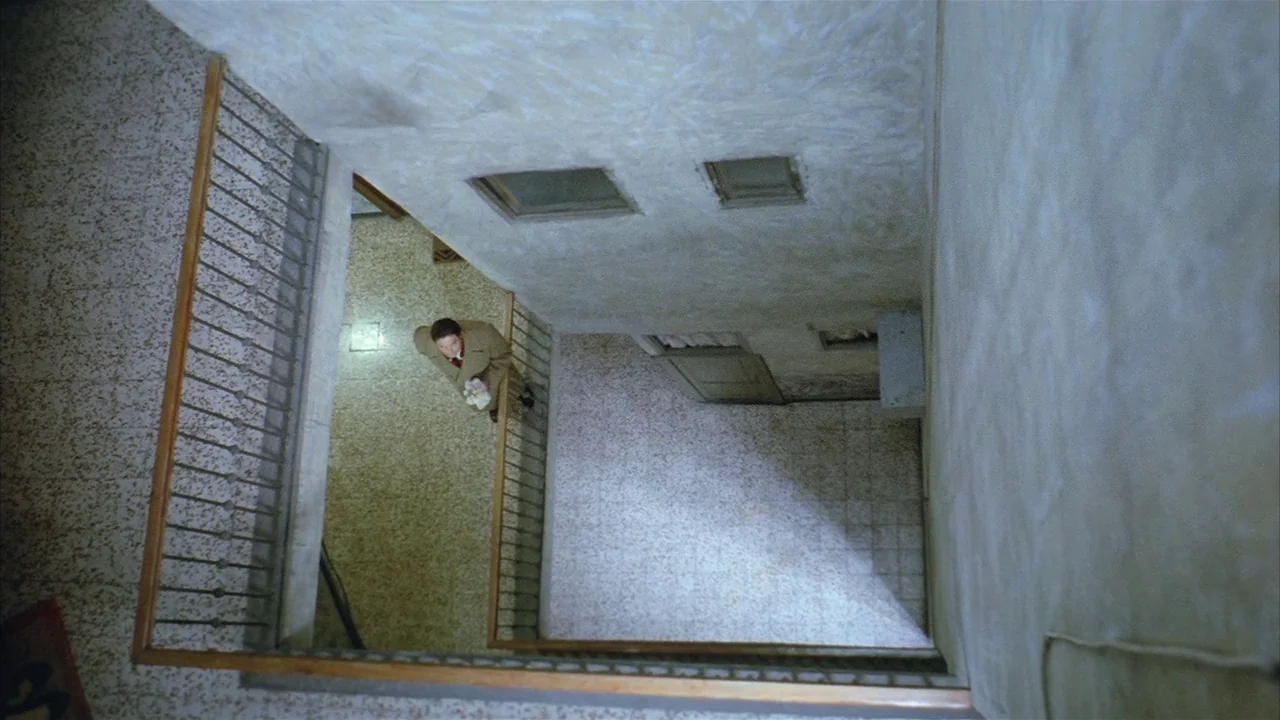Death Occurred Last Night (1970) movie still: wide — A vertical view looks down an austere, multi-story apartment stairwell with speckled tile…