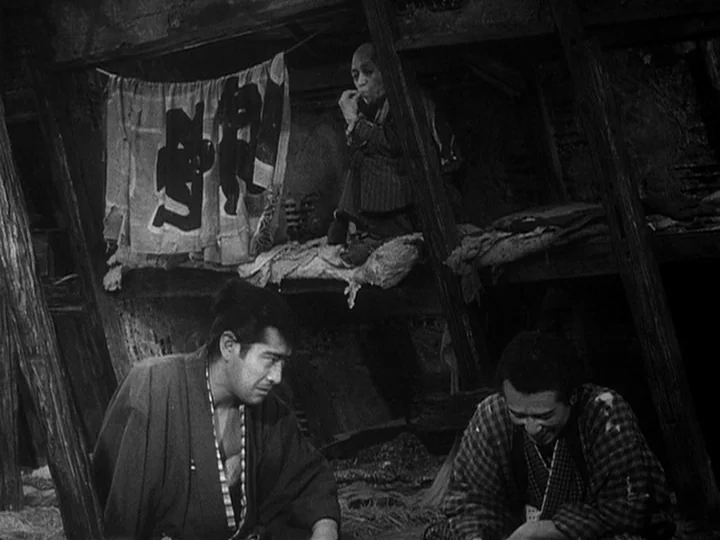The Lower Depths (1957) movie still: medium — A dark wooden structure contains cramped sleeping bunks and hanging fabric banners. Two men sit on the…