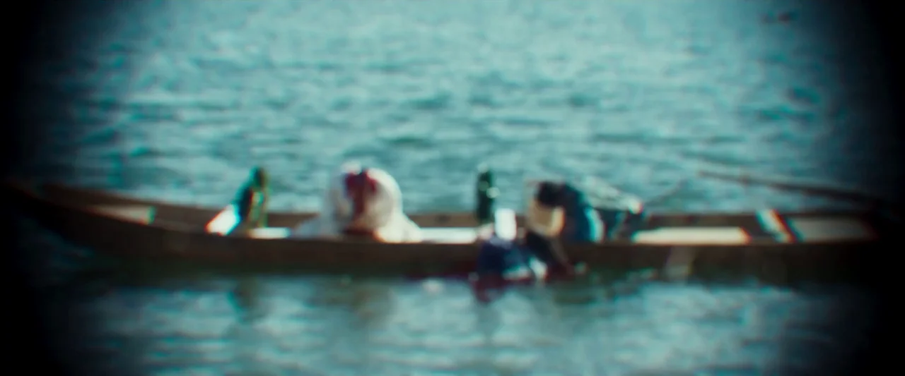 Kubi (2023) movie still: wide — A long wooden boat sits on the rippling surface of a large body of water. The subject is out of focus, appearing as…