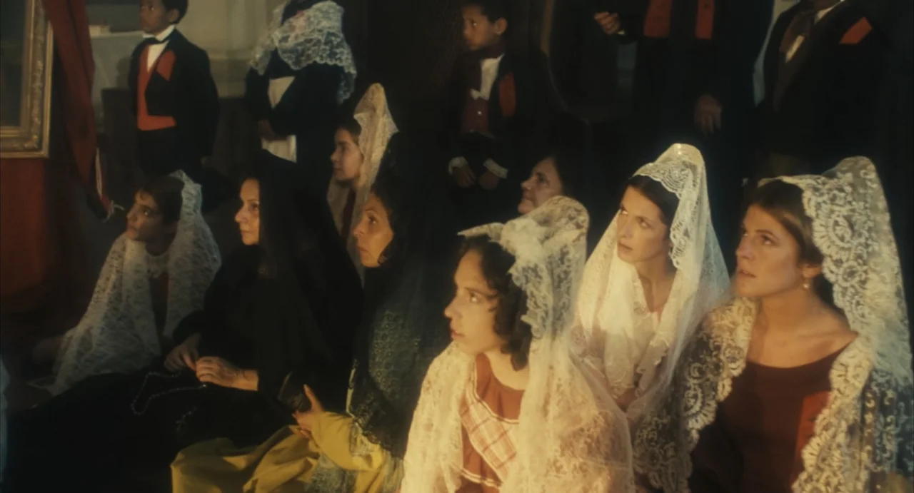 Camila (1984) movie still: medium — A dimly lit indoor space with several women and children gathered in a row. They wear lace veils and traditional…