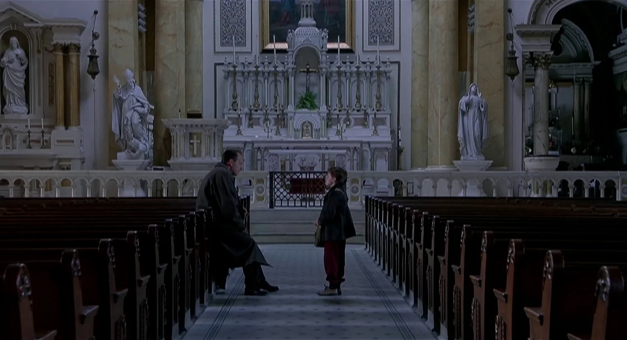 The Sixth Sense (1999) movie still: wide — A quiet cathedral nave features symmetrical wooden pews flanking a tiled central aisle. An adult man sits…