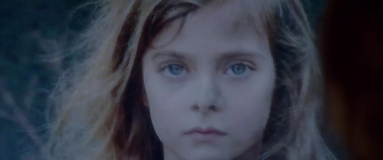 Elle (2016) movie still: closeup — A child stands outdoors in dim, cool-toned light with a blurred background. The subject faces the camera directly…