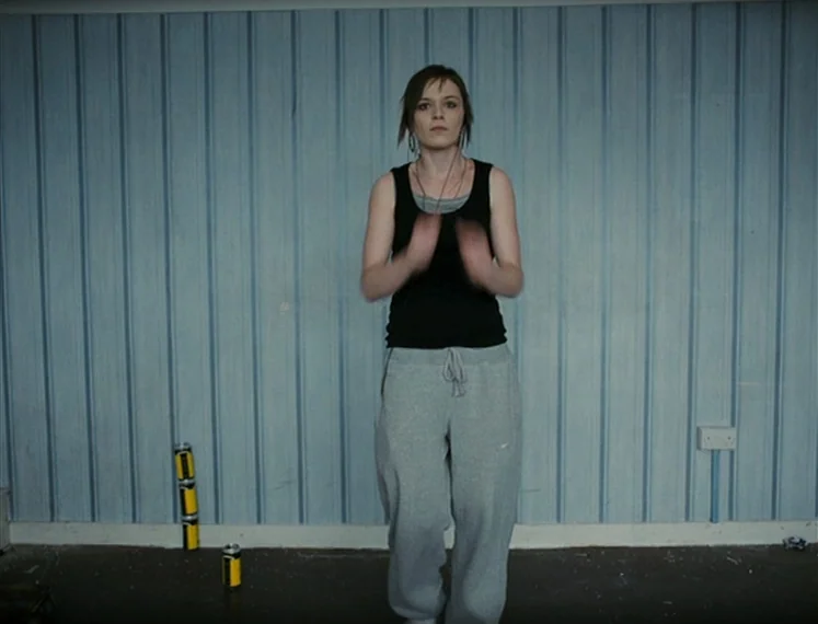 Fish Tank (2009) movie still: fullbody — A stark room features pale blue vertically paneled walls and a plain floor. A young woman stands in the…