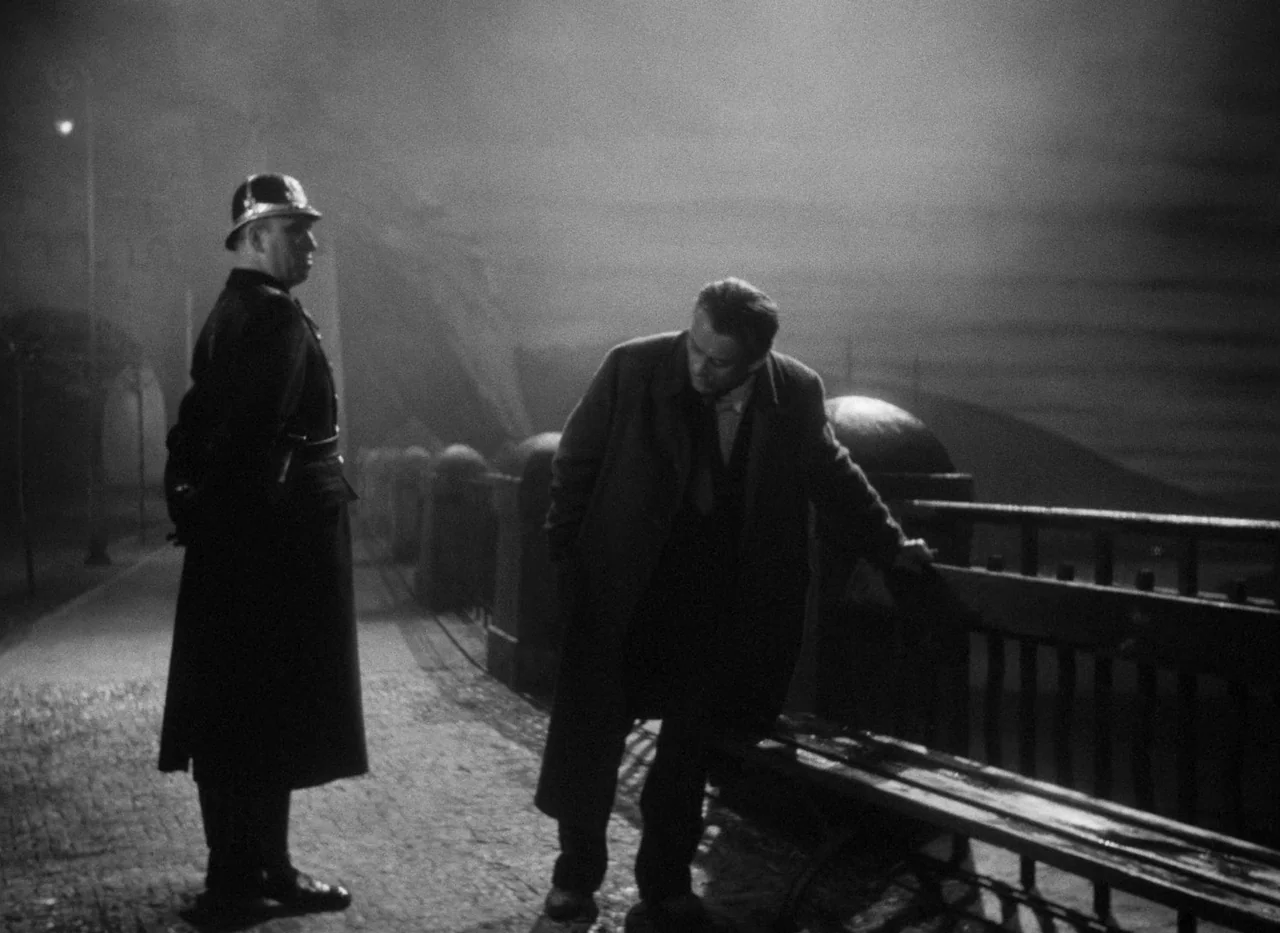 Krakatit (1948) movie still: medium — A dark, misty stone bridge at night lit by a single overhead lamp. A man in a coat leans against a bench while…