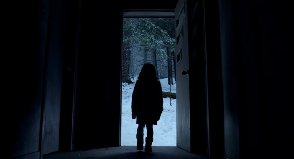 Mama (2013) movie still: fullbody — A dark indoor space looks out through an open door into a snowy forest. A young child stands in the center of…