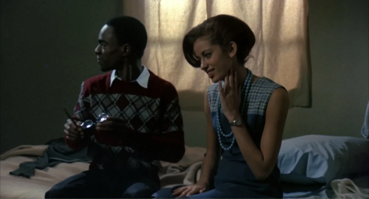 Cooley High (1975) movie still: medium — A furnished interior bedroom with a window covered by a light-colored curtain. A man and a woman sit side…