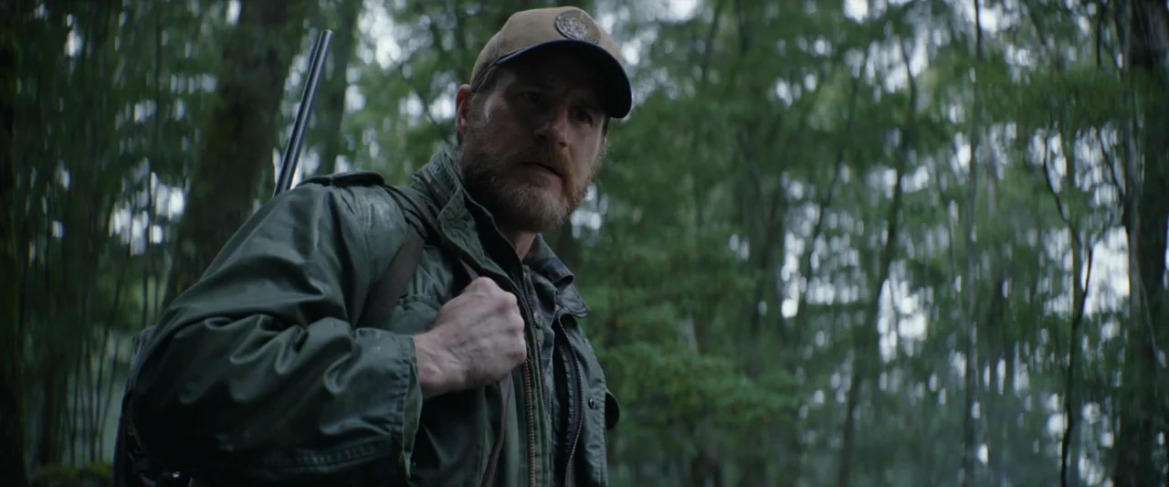 Wolf Man (2025) movie still: medium — A man walks through a dense woodland with tall trunks and blurred foliage behind. He wears an olive green…