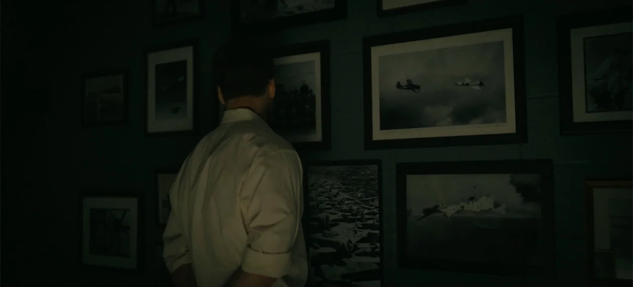 Devotion (2022) movie still: medium — A dark office wall is covered with multiple framed black and white aerial photographs. A man in a…