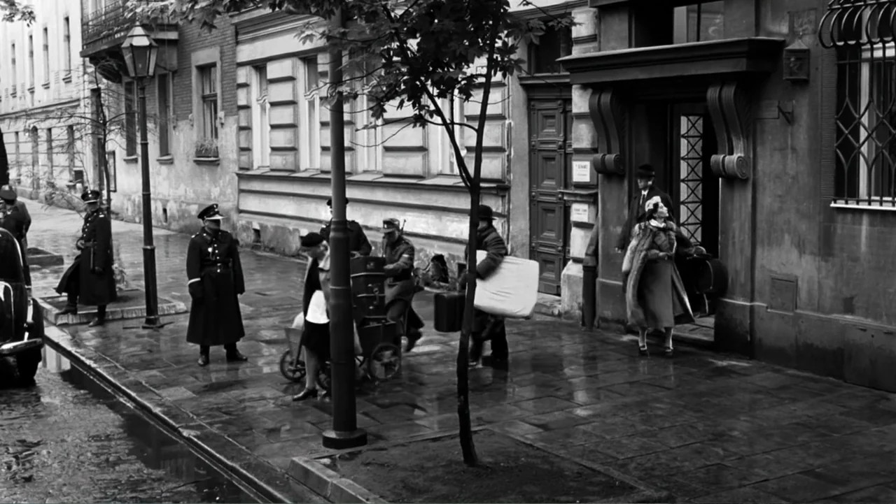 Schindler's List (1993) movie still: wide — A wet urban sidewalk in front of a stone building with uniformed armed guards. Civilians carrying…