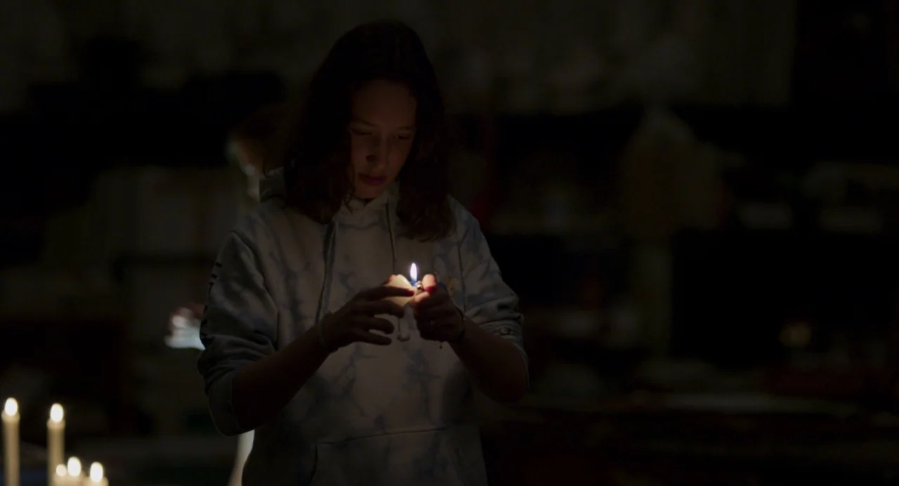 Zombi Child (2019) movie still: medium — A dark space features a teenager standing amidst several small candles on a surface. The young person in a…