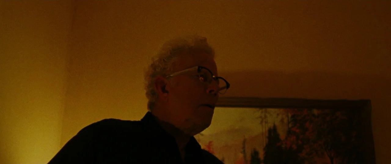 The Old Man & The Gun (2018) movie still: medium — An older man stands in a dimly lit room with an orange-hued light source. A framed landscape…