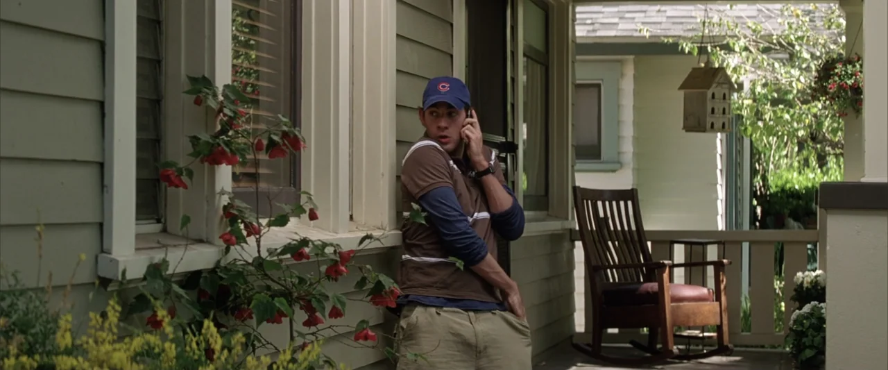 License to Wed (2007) movie still: medium — A wooden house porch features green siding, a rocking chair, and potted red flowers. A young man stands…