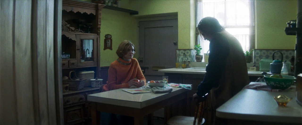 Starve Acre (2023) movie still: medium — A 1970s-style room with pale green walls and wooden cabinets around a table. A woman sits holding an orange…