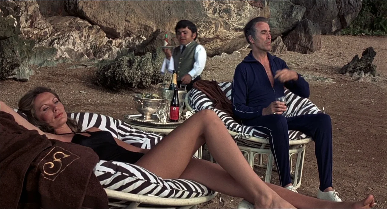 The Man With the Golden Gun (1974) movie still: wide — A rocky beach cove features striped wicker chairs placed on coarse sand. Three people lounge…