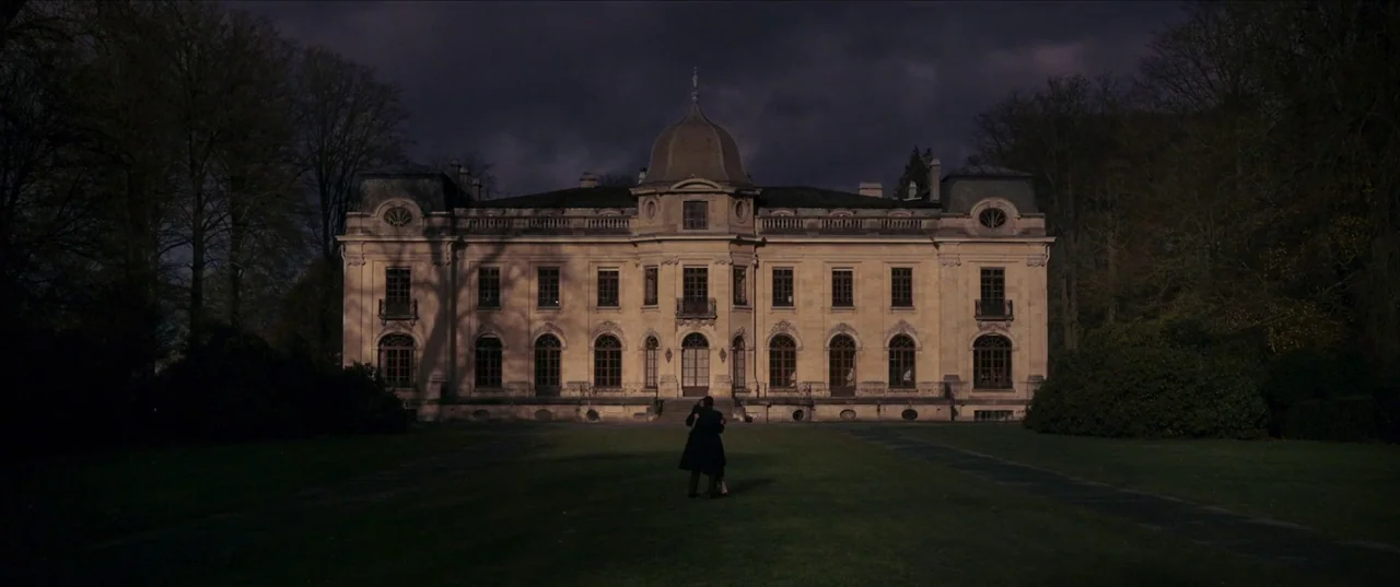 Earwig (2022) movie still: establishing — A large stone chateau with a central dome stands against a dark, cloudy sky. Two figures stand embracing…