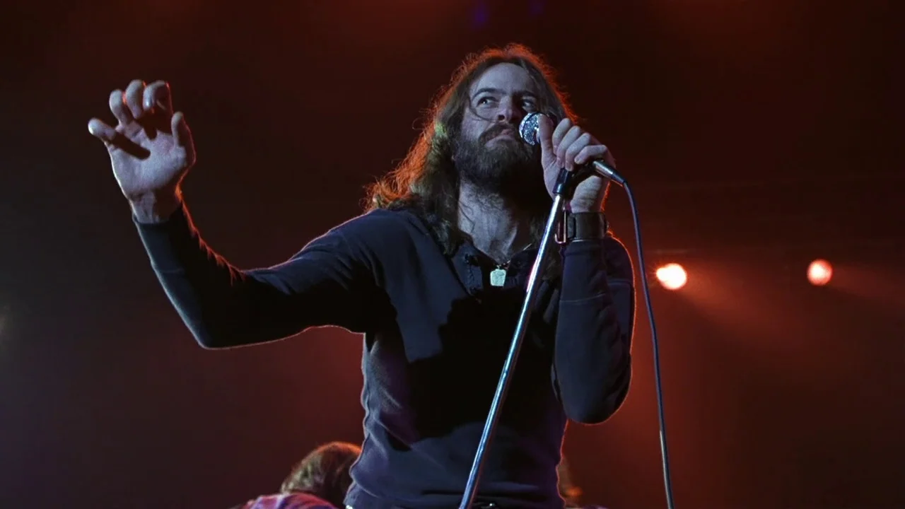 Almost Famous (2000) movie still: medium — A dimly lit stage features a bearded singer performing under intense red lights. He stands before a…